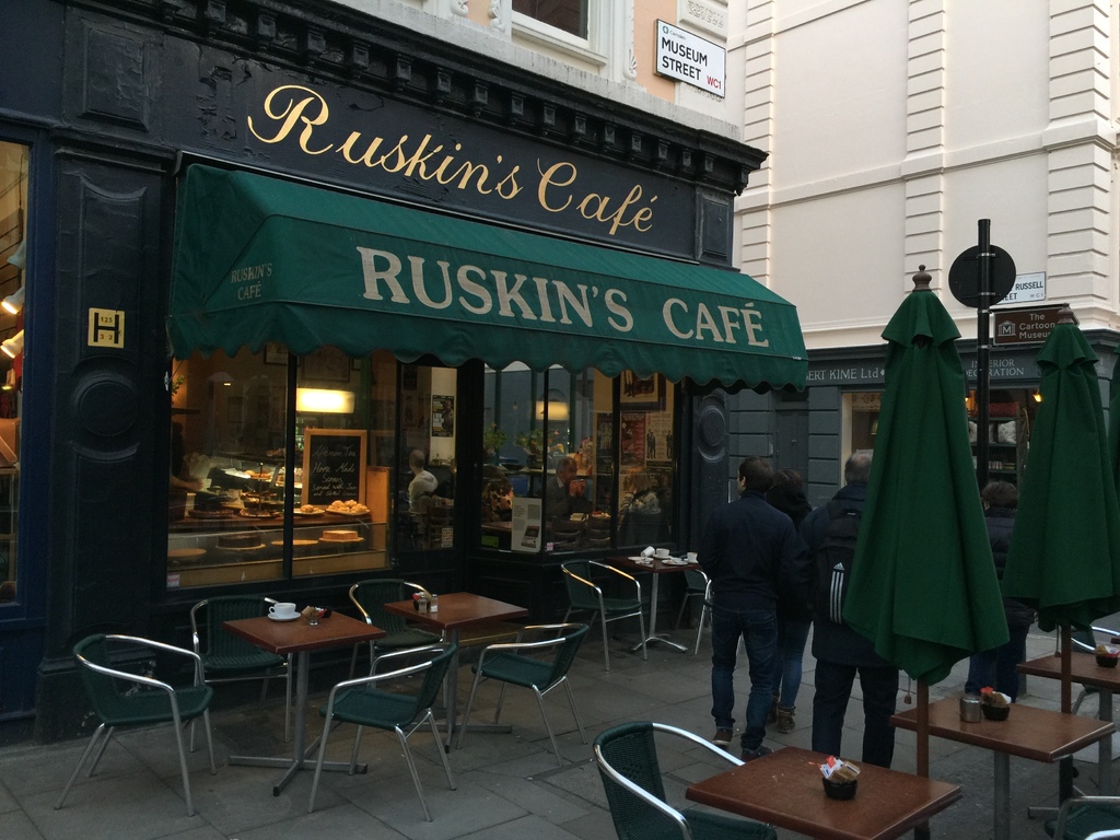 Eine Gruppe von Menschen vor einem Cafe mit einer Tafel, auf der "Ruskin's Cafe" steht.