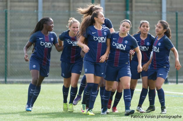 Gruppe junger Frauen, die auf einem Rasenfußballfeld spielen, mit Maschendrahtzaun und einer Wand im Hintergrund, Text in der rechten unteren Ecke lautet "Paris Saint-Germain Frauenfußball".