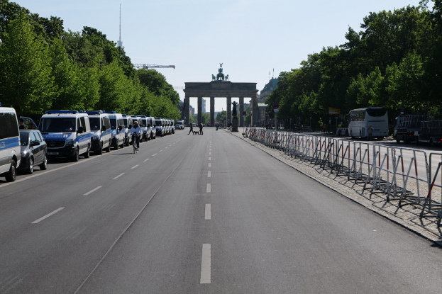 Eine lange Reihe von Polizeiwagen, die auf der Seite einer Straße vor dem Brandenburger Tor in Berlin, Deutschland, geparkt sind, mit Menschen, die Fahrräder fahren und auf der Straße stehen, Barrieren, Bäumen und einem Bogen mit Statuen im Hintergrund.