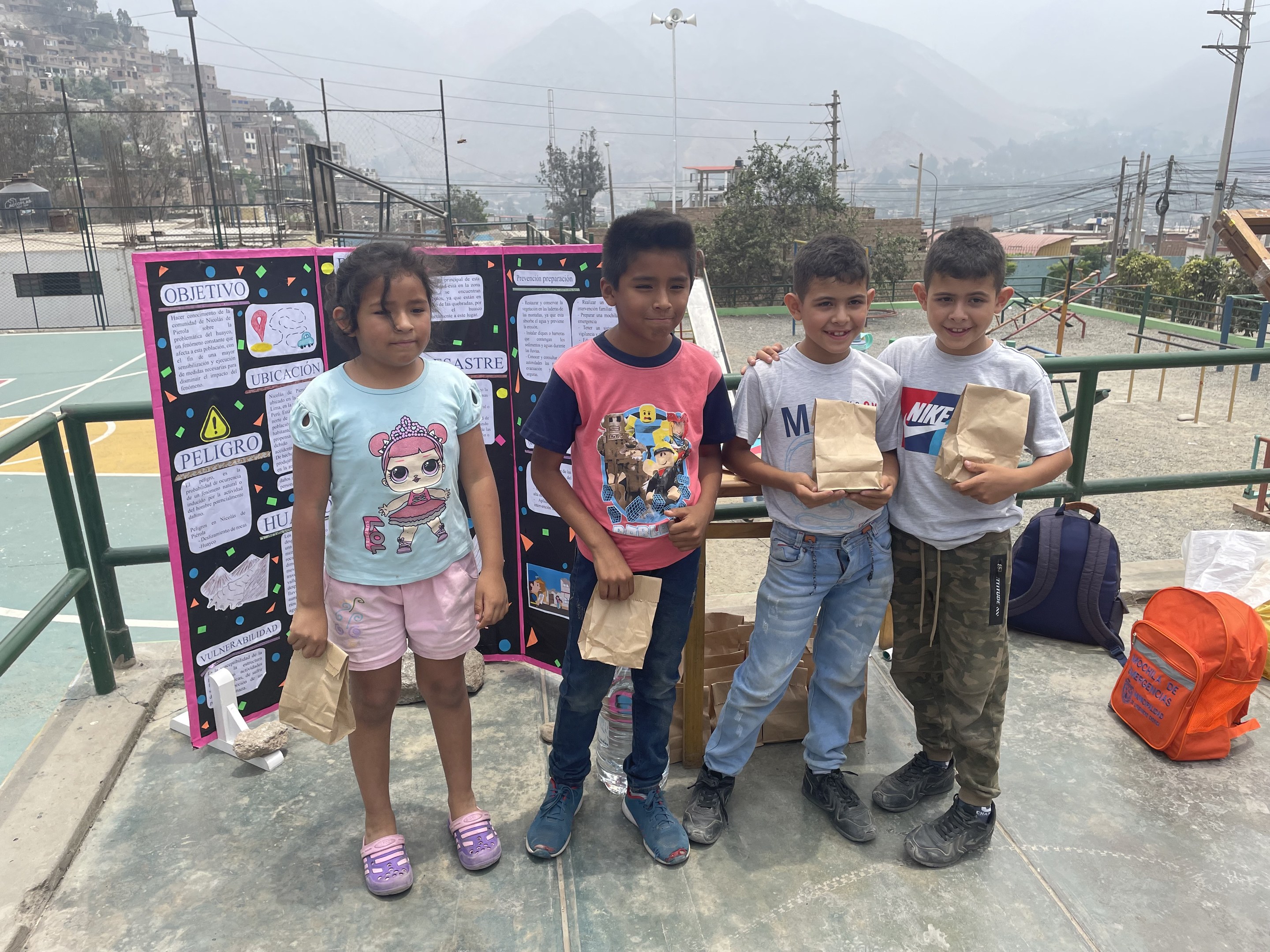 Gruppe von Kindern auf einem Basketballfeld mit Papiertüten in den Händen, Tüten und Gegenstände rechts und eine Tafel mit Text links, vor einer Outdoor-Background mit Zaun, Pfosten, Straßenlaternen, Bäumen, Gebäuden, Hügeln und Himmel.