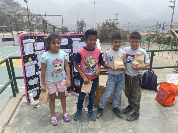 Gruppe von Kindern auf einem Basketballfeld mit Papiertüten in den Händen, Tüten und Gegenstände rechts und eine Tafel mit Text links, vor einer Outdoor-Background mit Zaun, Pfosten, Straßenlaternen, Bäumen, Gebäuden, Hügeln und Himmel.