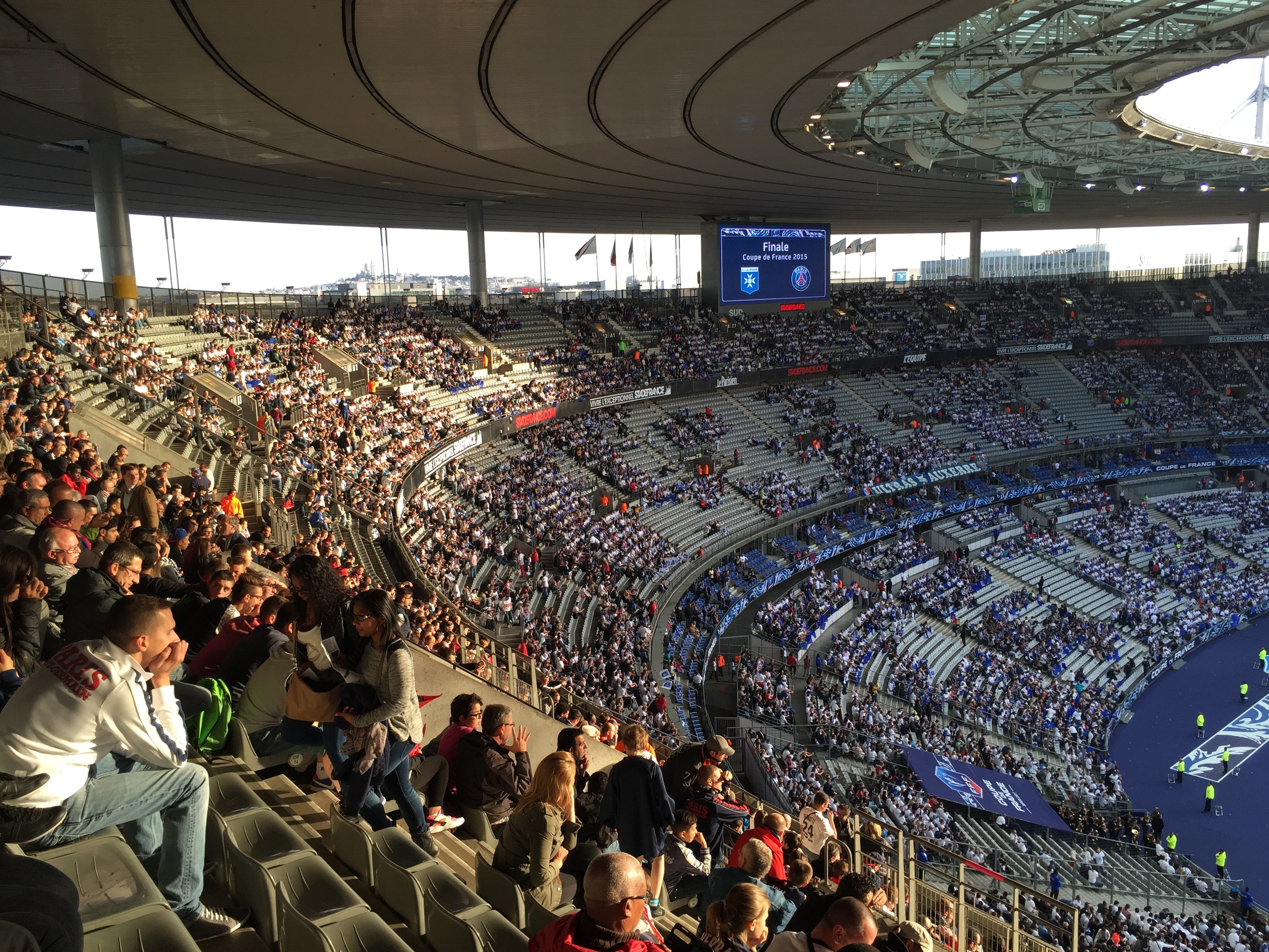 Große Zuschauermenge in einem Stadion bei einem Fußballspiel mit einer Bühne, Fahnen, Stangen und einem Bildschirm im Hintergrund.