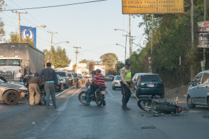 Eine Gruppe von Menschen steht um ein verunglücktes Motorrad auf der Seite der Straße mit mehreren Fahrzeugen, darunter ein Lastwagen, und einer Hintergrund von Bäumen, Pfählen, Lampen und Schildern unter einem Himmel.