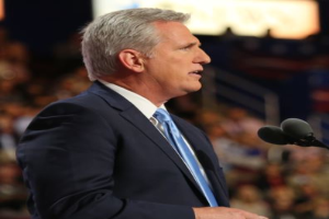 Ein Mann im Anzug und Krawatte steht an einem Podium mit zwei Mikrofonen, umgeben von einem unscharfen Hintergrund, und sieht ernst aus.