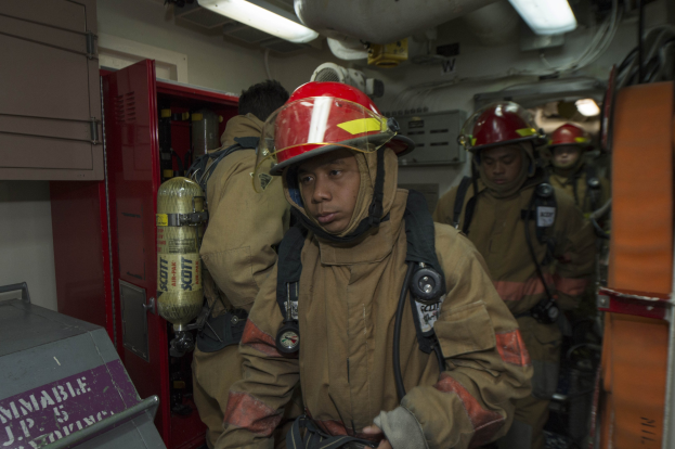 Feuerwehrleute in Uniform und Helmen, einer hält einen Feuerlöscher, mit einem metallischen Objekt links und Hintergrundelementen einschließlich Lichter und Rohre.