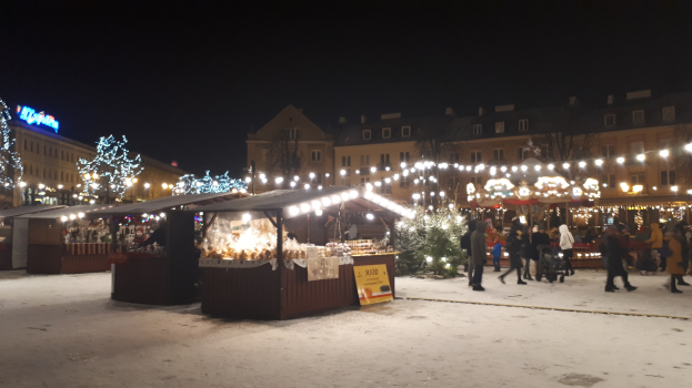 Ein belebter Weihnachtsmarkt an einem verschneiten Abend mit Menschen, Buden mit Lichtern, Pflanzen, Bäumen, Gebäuden, Schildern und einem bewölkten Himmel.