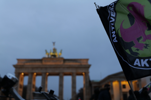Gruppe von Menschen demonstrieren vor dem Brandenburger Tor mit einer Fahnenstange, die ein Logo und Text trägt.