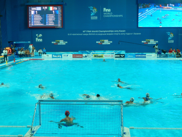 Gruppe von Athleten, die Wasserball in einem Schwimmbecken während der FINA-Weltmeisterschaften 2016 in Kazan spielen, mit einem Netz, das das Becken teilt, und Zuschauern, die links sitzen.