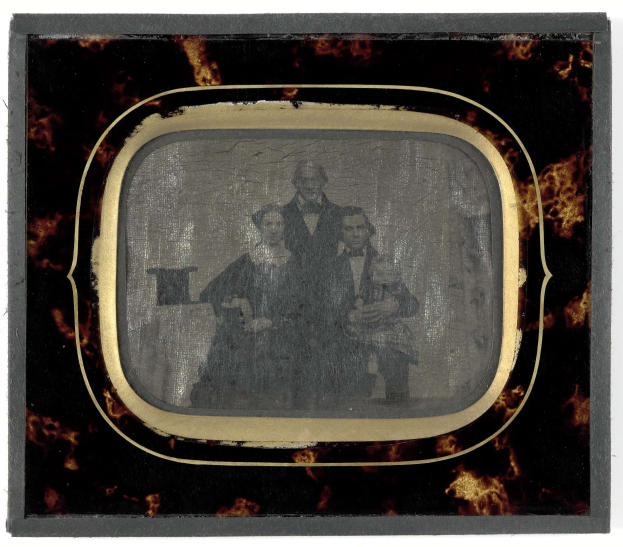 Schwarz-weißes Foto eines Mannes in Anzug und Krawatte und einer Frau in einem unbekannten Outfit, die vor einem alten Fernseher stehen und ernste Gesichter haben.