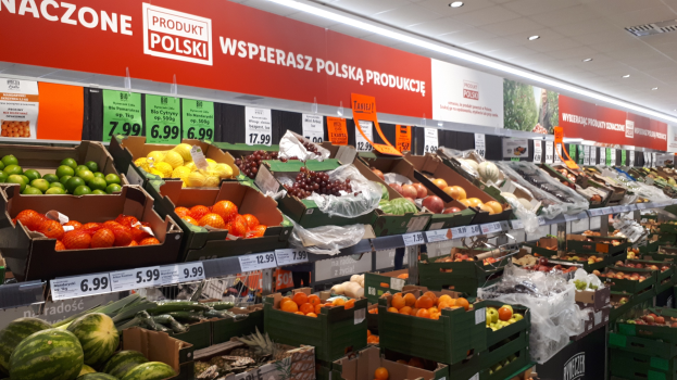 Ladeninneres mit verschiedenen Früchten und Gemüsen in Kartons mit Preisschildern, Werbetafeln an den Wänden und Deckenbeleuchtung.