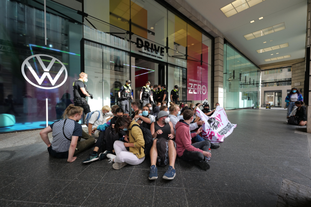 Eine Gruppe von Menschen sitzt vor einer Volkswagen-Vertretung mit Glaswänden auf dem Boden und hält eine Schilder mit Text, während Lichter an der Decke leuchten, was auf eine Protestaktion hindeutet.