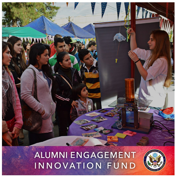 Gruppe von Menschen um einen Tisch versammelt, der ein "Alumni Engagement Innovation Fund"-Schild zeigt, mit Gegenständen auf dem Tisch und Zelten im Hintergrund unter einem klaren blauen Himmel.