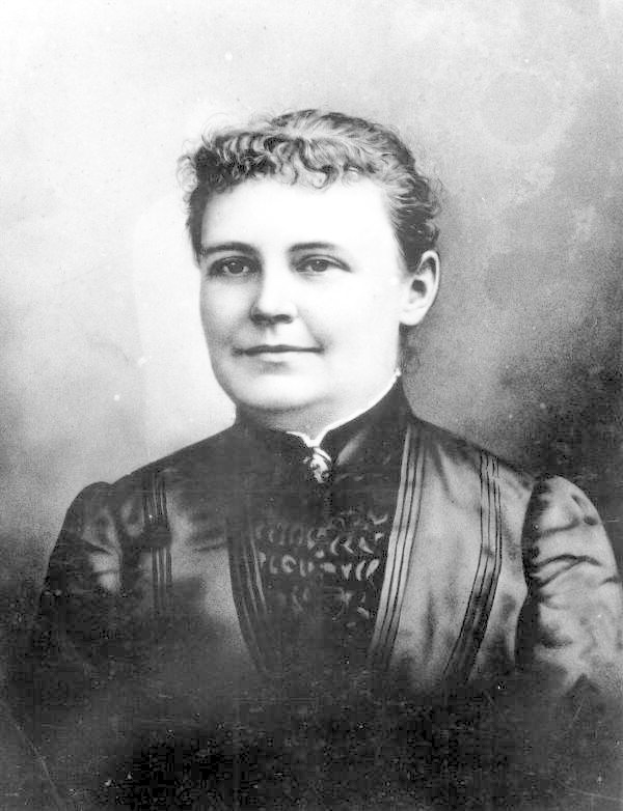 Schwarz-weißes Foto einer Frau in einem Kleid, die mit gefalteten Händen steht und einen entschlossenen Gesichtsausdruck hat.