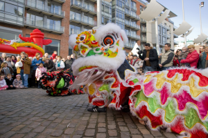 Eine Löwentanzvorstellung während eines chinesischen Neujahrsfestes in Amsterdam, mit einer Zuschauermenge und Menschen, die den Moment mit Kameras festhalten, vor einem Hintergrund von Gebäuden und einem klaren blauen Himmel.