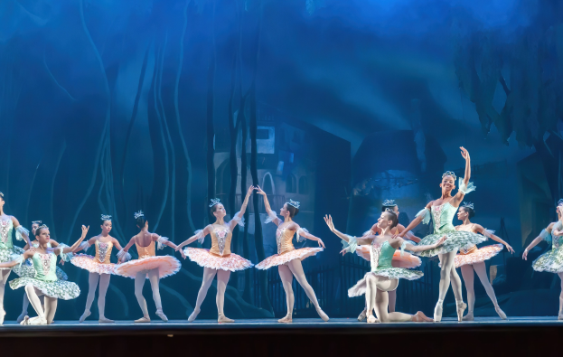 Eine Gruppe von Balletttänzern in bunten Kostümen, die *Der Nussknacker* auf der Bühne des Royal Opera House tanzen, bewegen sich synchronisiert gegen einen blauen Hintergrund.