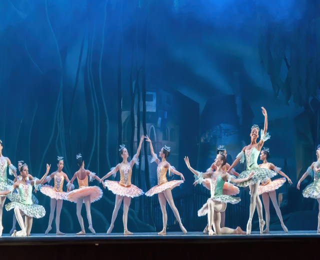 Eine Gruppe von Balletttänzern in bunten Kostümen, die *Der Nussknacker* auf der Bühne des Royal Opera House tanzen, bewegen sich synchronisiert gegen einen blauen Hintergrund.
