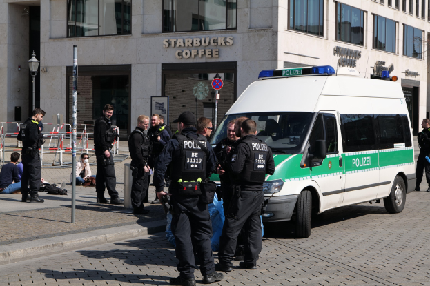Eine Gruppe von Polizisten vor einem Starbucks-Café mit einem Van auf der rechten Seite und einigen Menschen auf der linken Seite, mit einem Gebäude, einem Schild, einem Laternenpfahl und einem Zaun im Hintergrund.