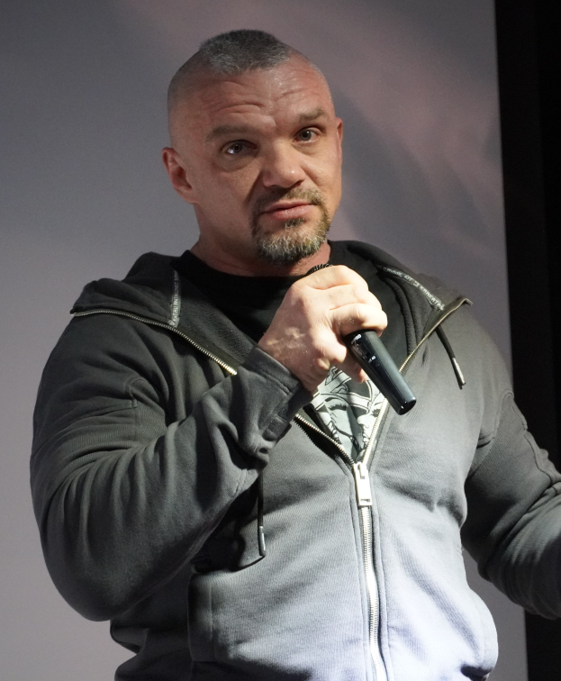 Ein Mann in einer schwarzen Kapuzenjacke und einem schwarzen Anzug, der ein weißes Hemd trägt, hält ein Mikrofon und steht selbstbewusst vor einer Wand, was darauf hindeutet, dass er ein Komiker ist.