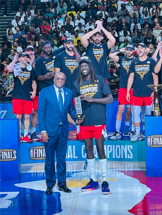 Eine Gruppe von Männern auf einem Basketballfeld feiert mit Trophäen, während Menschen im Hintergrund sitzen und zwei Trophäen rechts auf Podesten stehen.