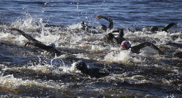 Eine Gruppe von Menschen in schwarzen Neoprenanzügen und Helmen, die synchron im Triathlon schwimmen, mit reflektierender Sonne auf dem Wasser.