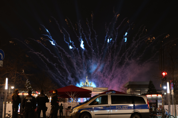Ein Polizeiwagen steht vor einer Menschenmenge, umgeben von Fahrrädern, Zelten, Laternenmasten, Schildern, Ampeln, Bäumen, Gebäuden und einer Statue im Hintergrund, unter einem von Feuerwerk erhellten Himmel.