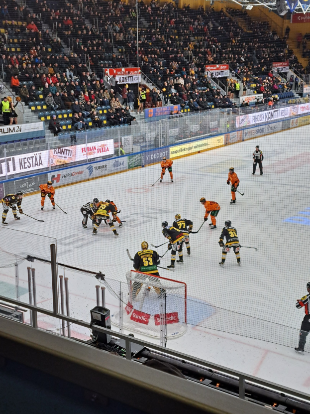 Eine Gruppe von Menschen, die Hockey auf einem Eisring spielen, umgeben von einem Zaun, mit Zuschauern im Hintergrund und beleuchteter Stadionbeleuchtung oben.