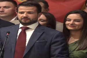 Ein Mann im Anzug spricht in ein Mikrofon an einem Podium, mit einem Banner dahinter und einer aufmerksamen Menge davor.