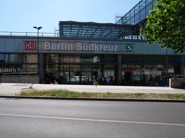 Große gläserne Berliner Südkreuz-Bahnhof mit sichtbarem Text, umgeben von Straßeninfrastruktur, Fahrzeugen, Füpßen, Fahrrädern, Bäumen und einem klaren Himmel.