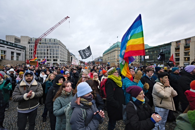 Große Gruppe von Menschen bei der LGBTQ+-Rechtsdemo in Berlin, mit Fahnen und Plakaten, vor einem Gebäude, mit Kränen und bewölktem Himmel im Hintergrund.