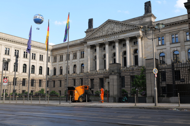 Großes modernes Gebäude mit zahlreichen Fenstern, als Bundestag in Berlin, Deutschland identifiziert, umgeben von Straßeninfrastruktur, Fußgängern, Flaggen und Bäumen unter einem bewölkten Himmel.