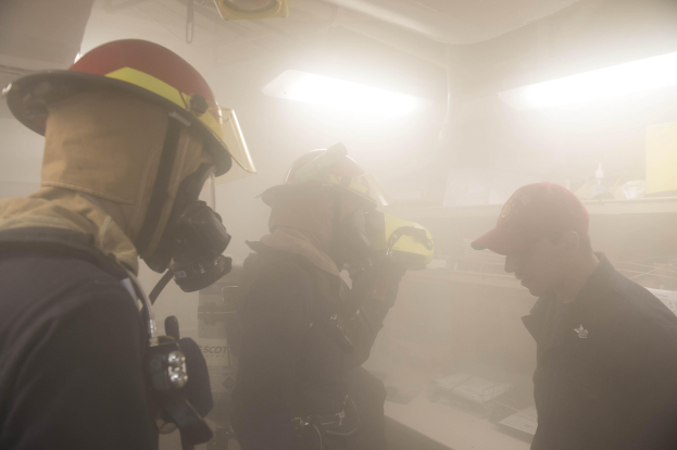 Feuerwehrleute mit Helmen und Rauch vor dem Mund, einer hält eine Kamera, während einer Trainingsrunde mit Hintergrundbeleuchtung und Gegenständen.