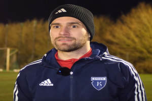 Ein Mann in einer blauen Jacke und Mütze steht auf einem Fußballfeld während eines Trainings, mit Bäumen und einem Tor im Hintergrund.