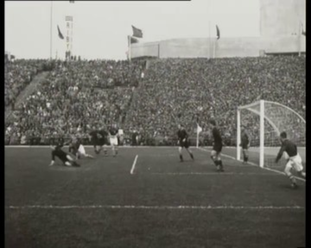 Schwarze-weißes Foto des WM-Finals 1958 zwischen Manchester United und Liverpool, das Spieler auf dem Feld, ein Torpfosten auf der rechten Seite, Zuschauer in den Rängen und Fahnen im Hintergrund zeigt.