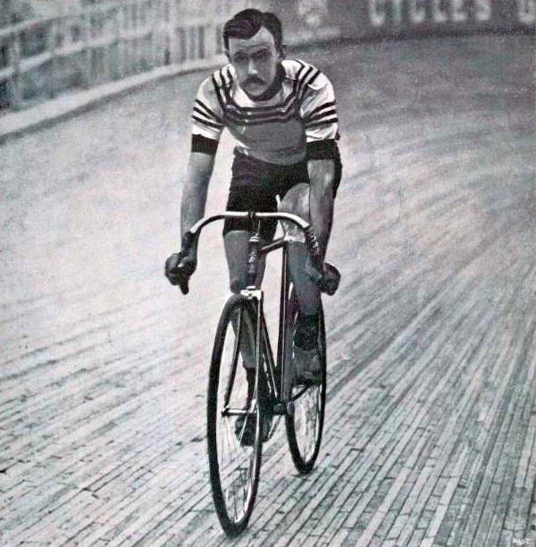 Schwarz-weißes Foto eines Mannes, der auf einer Bahn radelt, mit einem Zaun und einer Tafel im Hintergrund.