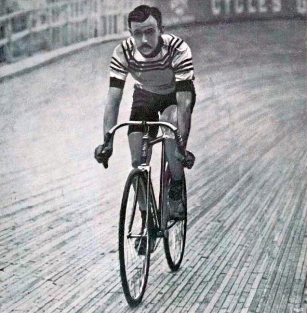 Schwarz-weißes Foto eines Mannes, der auf einer Bahn radelt, mit einem Zaun und einer Tafel im Hintergrund.