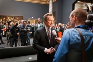Ein Mann im Anzug und Krawatte spricht mit einem Reporter vor einem Publikum, einige halten Kameras, mit einem Sofa im Vordergrund und einer Wand mit einem Foto, Lampen und einer Säule im Hintergrund, während er ein Mikrofon hält.