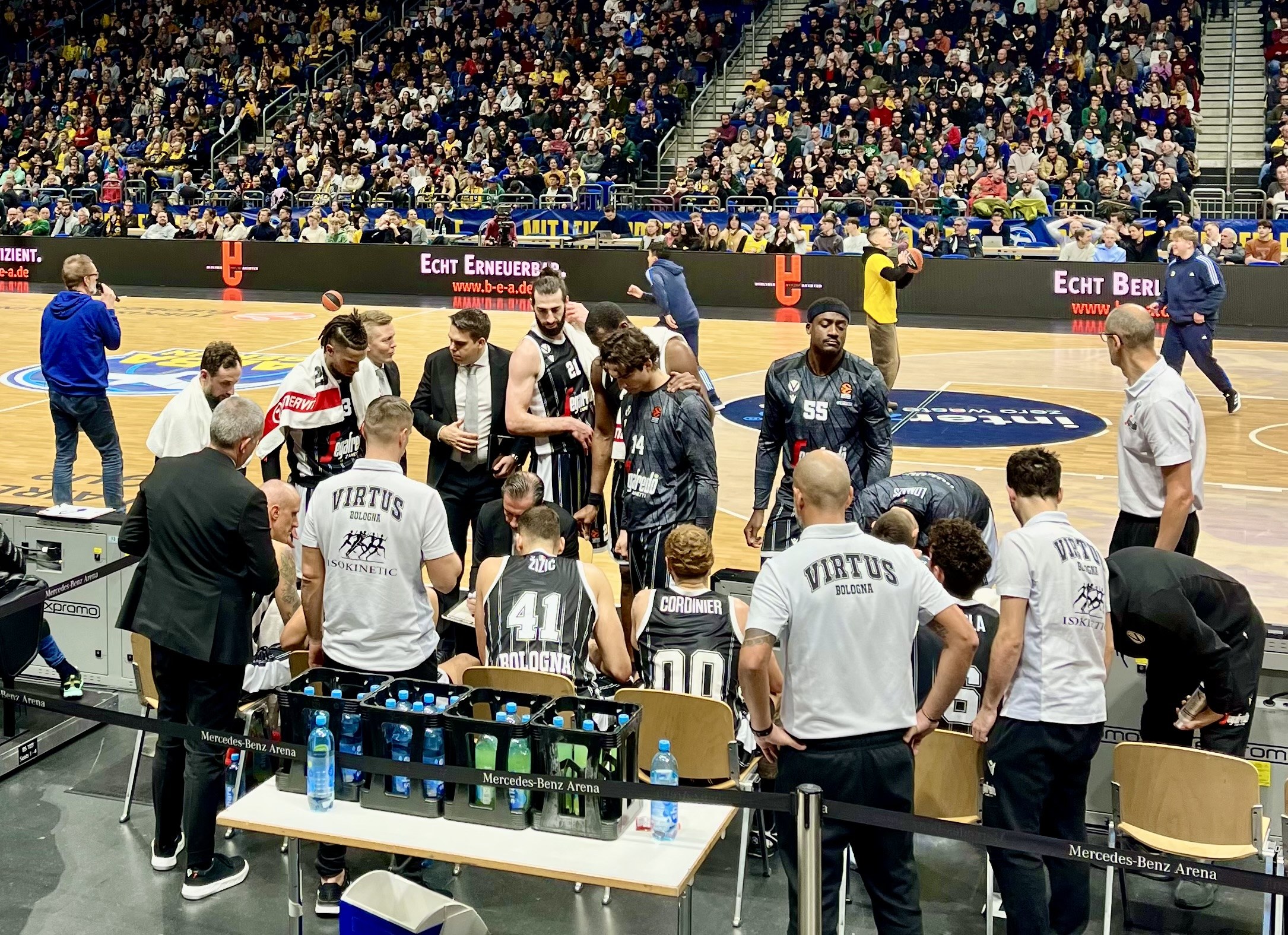 Eine Gruppe von Männern auf einem Basketballfeld während eines Spiels zwischen Virtus Berlin und Echt Berlin, mit Zuschauern auf Sitzplätzen und Stehplätzen, einem Fotografen und Tafeln mit Teamnamen im Hintergrund.