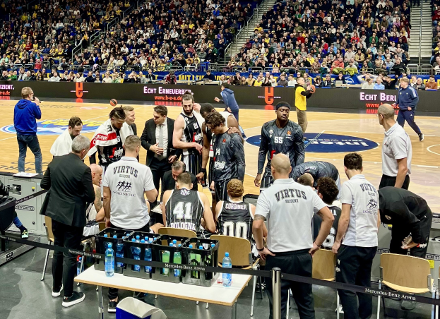 Eine Gruppe von Männern auf einem Basketballfeld während eines Spiels zwischen Virtus Berlin und Echt Berlin, mit Zuschauern auf Sitzplätzen und Stehplätzen, einem Fotografen und Tafeln mit Teamnamen im Hintergrund.