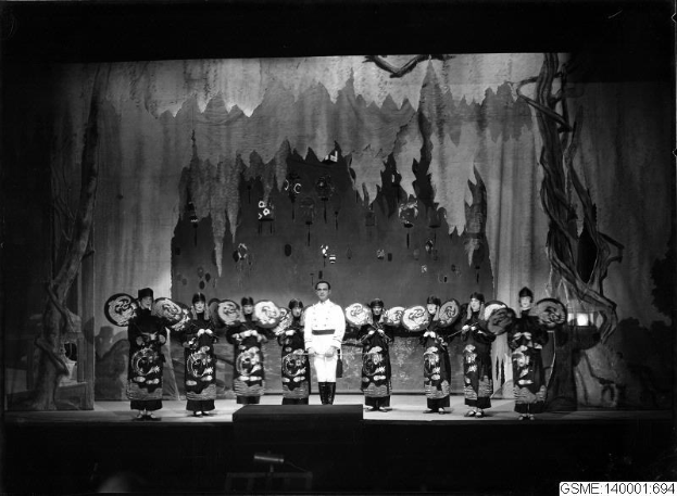 Schwarzes und weißes Foto von Darstellern in traditionellen japanischen Kimonos auf einer Bühne mit einer gemalten Kulisse, beschriftet mit 'Kimono-Show in Tokyo, Japan'.