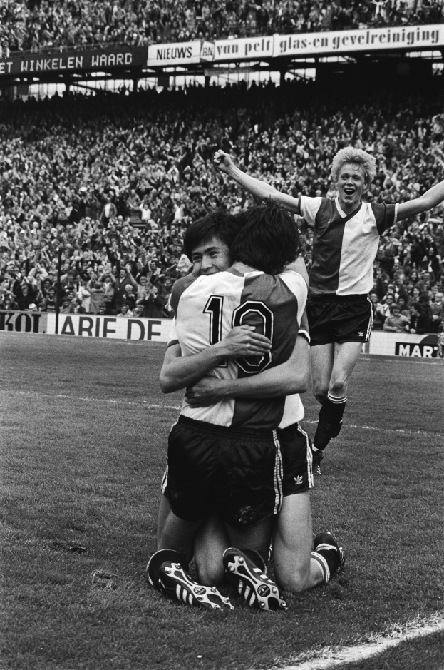 Drei Fußballspieler, die sich nach einem Tor auf dem Boden umarmen, vor einem jubelnden Publikum und schwenkenden Fahnen in einem schwarz-weißen Stadion.