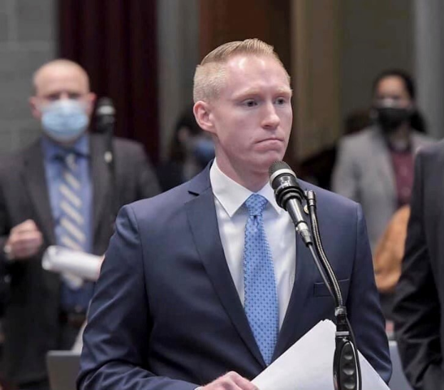 Ein Mann in Anzug und Krawatte spricht an einem Rednerpult mit Papieren, während er ein Mikrofon anspricht, während andere in der Nähe stehen.