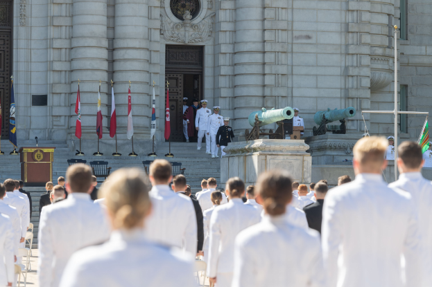 Gruppe von Menschen in weißen Marineuniformen, die vor einem Gebäude mit Säulen, Flaggen, einem Podium und Kanonen im Hintergrund stehen.