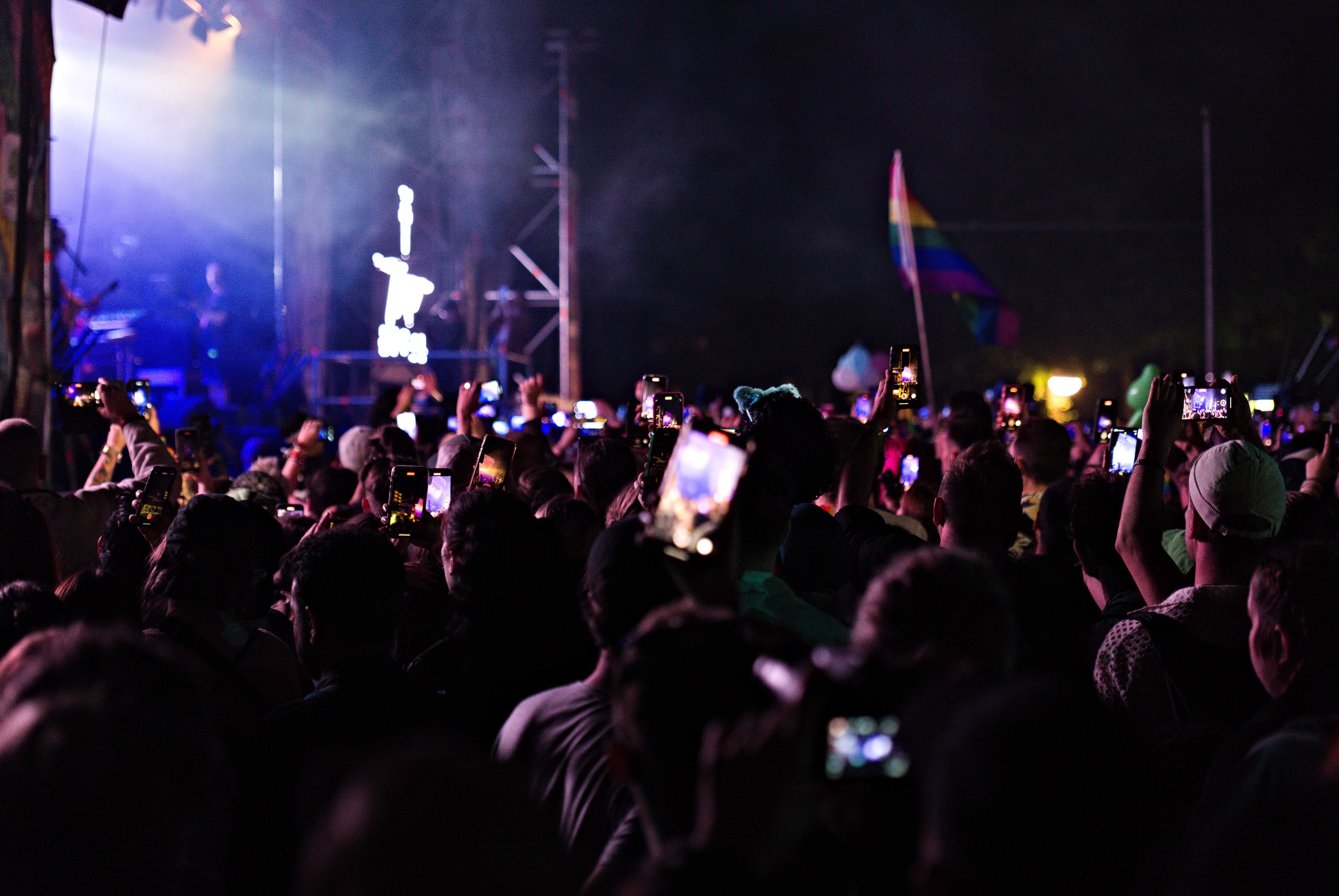 Eine Menge bei einem Konzert hält beleuchtete Handys hoch, während Musiker auf der Bühne spielen, umgeben von Pfählen und einer Fahne.