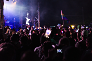 Eine Menge bei einem Konzert hält beleuchtete Handys hoch, während Musiker auf der Bühne spielen, umgeben von Pfählen und einer Fahne.