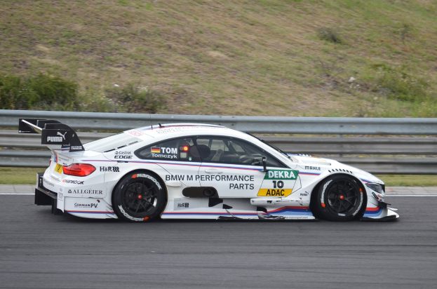 BMW M4 DTM-Rennwagen auf einer Rennstrecke mit Werbetext, umgeben von grünem Gras und einer Absperrung im Hintergrund.