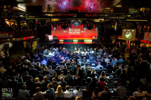 Eine große Menschenmenge sitzt auf Stühlen vor einer Bühne mit Bildschirmen und Bannern, die von Scheinwerfern beleuchtet wird und von Geländern umgeben ist, voller Erwartung auf den Beginn eines Online-Marketing-Rockstars-Events, mit einem Wasserzeichen in der linken unteren Ecke.