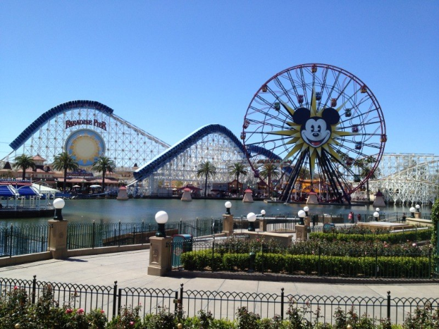 Luftaufnahme von Disney California Adventure mit seinem Riesenrad und Achterbahn, umgeben von Grünflächen und Wasser, unter einem klaren blauen Himmel.