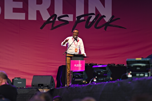Ein Mann in einem weißen Hemd und braunen Hosen steht auf einer Bühne und hält ein Mikrofon in der Hand, hinter ihm eine pinkfarbene Fahne mit der Aufschrift 'Berlin as Fuck' und sichtbare Lautsprecher.