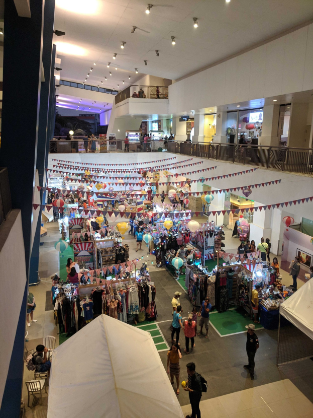Ein volles Einkaufszentrum mit Kleiderständen, Dekorationsflaggen, Ballons und Menschen, die zwischen Geländern, Säulen und Deckenlampen sitzen oder stehen, suggeriert einen Weihnachtsmarkt im Mall of Asia.