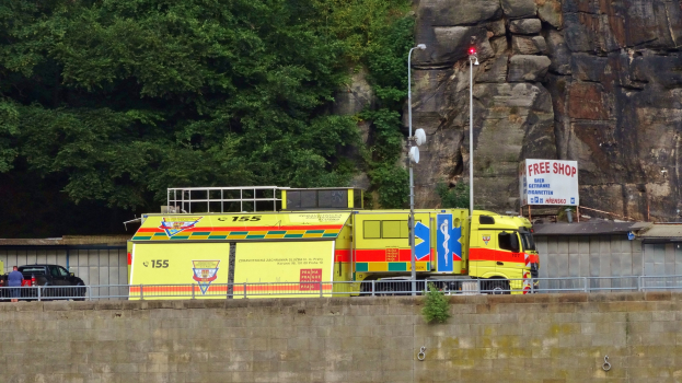 Ambulanz auf der Seite einer Straße neben einer Klippe geparkt, mit verschiedenen Objekten im Vordergrund und Bäumen und einem Auto im Hintergrund.